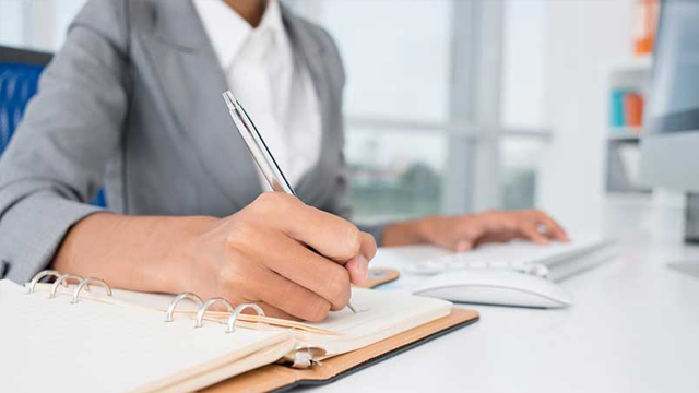 Imagem de uma mulher sentada à frente do computador. Ela digita algo no teclado com uma mão enquanto faz anotações num caderno com a outra.