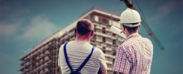 Foto de dois homens de costas olhando para um prédio em construção. Ao lado do prédio, um imenso guindaste. O homem da direita usa um capacete de segurança branco e aponta para o prédio. O da esquerda usa camiseta branca e macacão azul.
